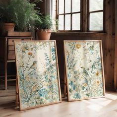 two framed paintings sitting on top of a wooden floor next to a potted plant