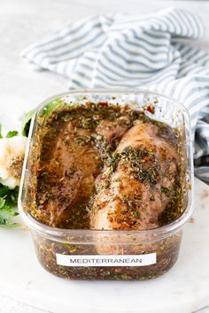 a plastic container filled with meat covered in seasoning next to some vegetables and herbs