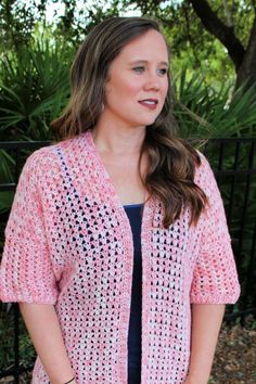 a woman standing in front of a fence wearing a pink crocheted cardigan