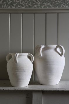 two white vases sitting on top of a shelf next to each other in front of a wall