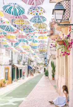 a woman sitting on the ground under umbrellas in an alleyway with potted plants