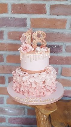 a three tiered cake with pink flowers and gold numbers on top, sitting against a brick wall