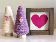 two small crocheted trees are next to a framed heart on a white table