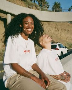 two young women sitting on the side of a road smiling at each other with their eyes closed