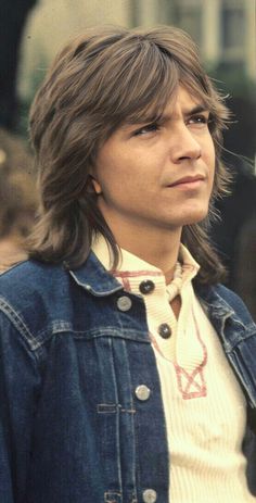 a man with long hair wearing a denim jacket and looking off to the side in front of a building