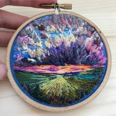 someone is holding up a colorful embroidery art work on a wooden hoop with a needle