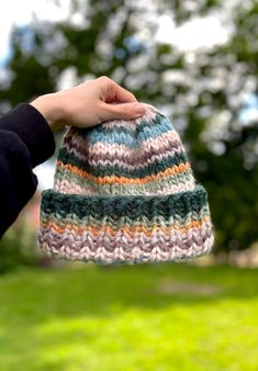 a hand holding a multicolored knitted hat in front of a green field