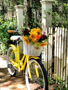 a yellow bicycle with flowers in the basket is parked next to a white picket fence