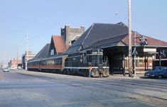 an old train station with cars parked on the tracks next to it and buildings in the background