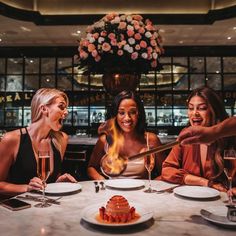 three women sitting at a table with wine glasses in front of them and a cake on the table