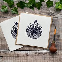 two cards with christmas ornaments on them sitting next to some greenery and an ink pen