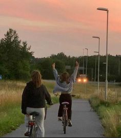 two women riding bikes on a path at dusk with their arms in the air and one woman raising her hand