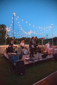 the band is playing on the stage in the yard at night with string lights strung across the fence