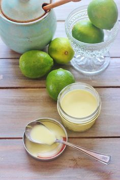 some limes are sitting on a wooden table next to two jars and spoons