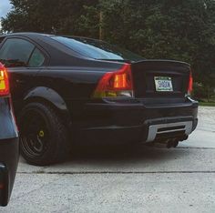 the back end of a black car parked in a parking lot next to some trees