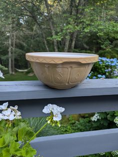 a bowl sitting on top of a wooden table next to white and blue flowers in front of trees