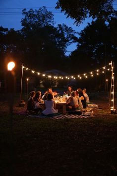 a group of people sitting around a table at night with lights strung from the trees