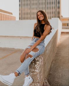 a woman sitting on a ledge with her legs crossed wearing ripped jeans and white sneakers