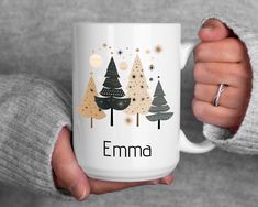 a person holding a white coffee mug with trees on it and the name emma