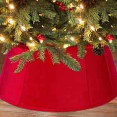 a christmas tree skirt with pine cones and red berries on the bottom, sitting in front of a lit christmas tree