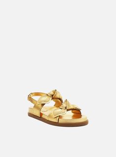 Golden leather flat sandal. The style features a rounded toe, a flat heel and an anatomically shaped insole with the brand name engraved. It has wide straps over the toes and upper foot, with an additional strap featuring topstitching and a knot, forming a large bow. There is a buckled strap around the heel. The sandal shows the foot. Why follow this trend? Set on a supportive footbed for all-day comfort, the Megan Sandal adds a playful touch with its large bows. It is perfect for laid-back occa Genuine Leather Sandals, Gold Flats, Leather Sandals Flat, Large Bow, Toe Sandals, Leather Buckle, Leather Flats, Wide Straps, Flat Sandals