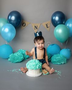 a baby sitting in front of a cake with blue frosting and balloons behind it