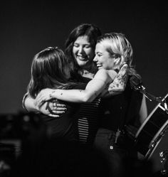 three women hug each other while standing in front of a drum set and microphones