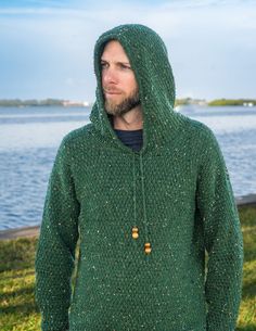 a man wearing a green hooded sweater standing next to the water with his hands in his pockets