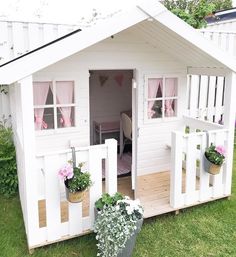 a small white house with pink curtains on the windows and flowers in potted plants