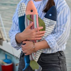 a woman holding a stuffed fish in her hands by the water's edge while wearing a blue striped shirt