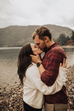 a man and woman standing next to each other on a rocky beach with mountains in the background