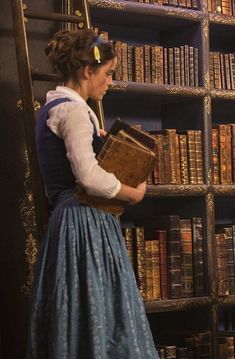 a woman in a blue dress is holding a book and looking at the bookshelf