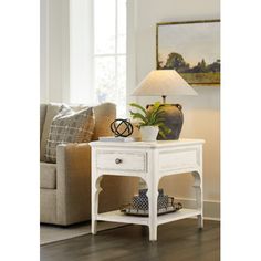 a white end table with a lamp on it in front of a couch and window