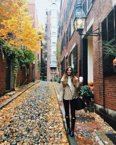 a woman is walking down an alleyway with leaves on the ground and buildings in the background