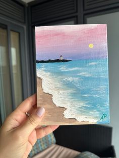 a person holding up a small painting in front of a window with the ocean on it