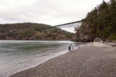 two people walking on the beach with a bridge in the background and trees lining the shore