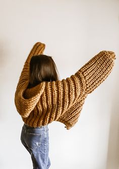 a woman wearing a sweater and jeans standing in front of a white wall with her arms wrapped around the back of her head