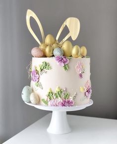 a cake decorated with flowers, eggs and bunny ears on a white pedestal in front of a gray wall