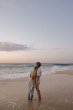 a man and woman standing on top of a sandy beach next to the ocean at sunset