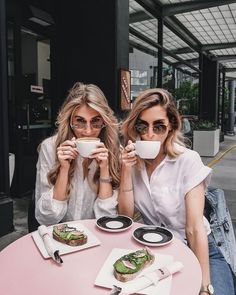 two women sitting at a pink table drinking coffee and eating sandwiches in front of them