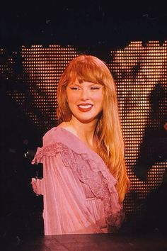 a woman with long red hair wearing a pink dress and smiling at the camera while standing in front of a screen