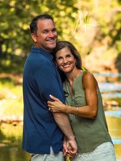 a man and woman hugging each other in front of a river with trees behind them