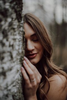 a woman with long hair leaning against a tree in the woods, holding her hand on it's face