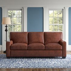 a brown leather couch sitting on top of a blue rug in front of two windows