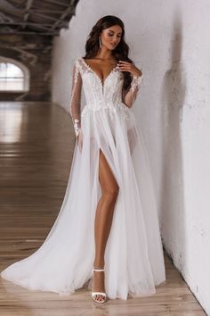 a woman wearing a long white dress with sheer sleeves and thigh high slits standing in an empty room