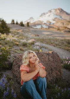 a woman sitting on top of a rock next to purple flowers and mountains in the background