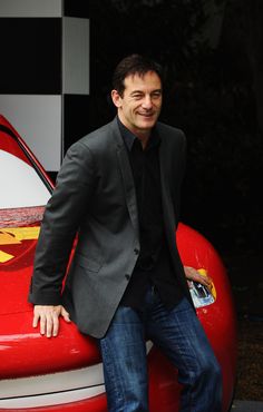 a man leaning on the hood of a red car with his hands in his pockets