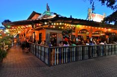 people are sitting at tables in front of an outdoor bar with lights on the roof