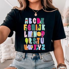 Colorful Alphabet Shirt, perfect for teachers of all levels, features a vibrant ABC design that's ideal for kindergarten and pre-K educators. Great gift idea for teachers!    Teachers, ditch the drab and embrace the fab! Is your current wardrobe stuck in a permanent state of "chalk dust disaster"?  Fear not, weary educators, for the sartorial savior has arrived!  Introducing the ABC Doodad, the most electrifying alphabet shirt this side of Sesame Street!  This vibrant masterpiece features a kaleidoscope of colors that'll have your little learners buzzing with excitement.  Imagine the look on their tiny faces as you waltz into class, a walking, talking alphabet symphony!  The ABC Doodad isn't just a shirt, it's a conversation starter, a learning tool disguised as delightful whimsy.  Don't l Cheap Novelty Shirt With Letter Print, Cute Teacher Shirts Shirts By Sarah, Alphabet Shirt, Pre K Teacher, Colorful Alphabet, Abc Design, Kindergarten Teacher Shirts, Kindergarten Teacher, Gift For Teacher