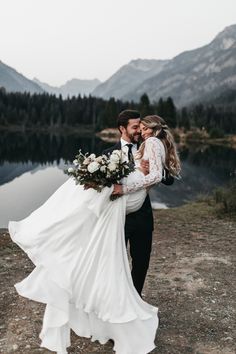 a bride and groom hugging each other in front of a lake with mountains behind them